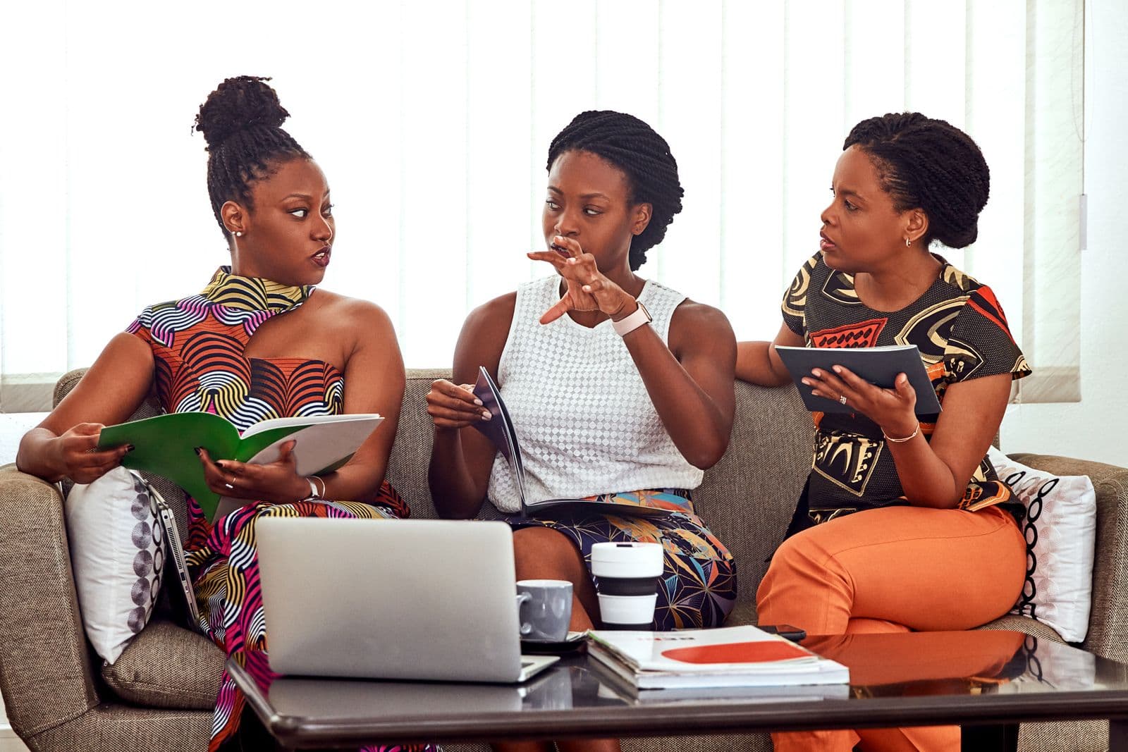 Three women in wax-print outfits talking around a laptop